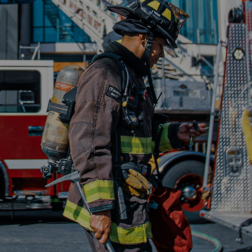 Veteran/Professional/Gov: Firefighter in protective gear standing in front of a fire truck, prepared for emergency response.